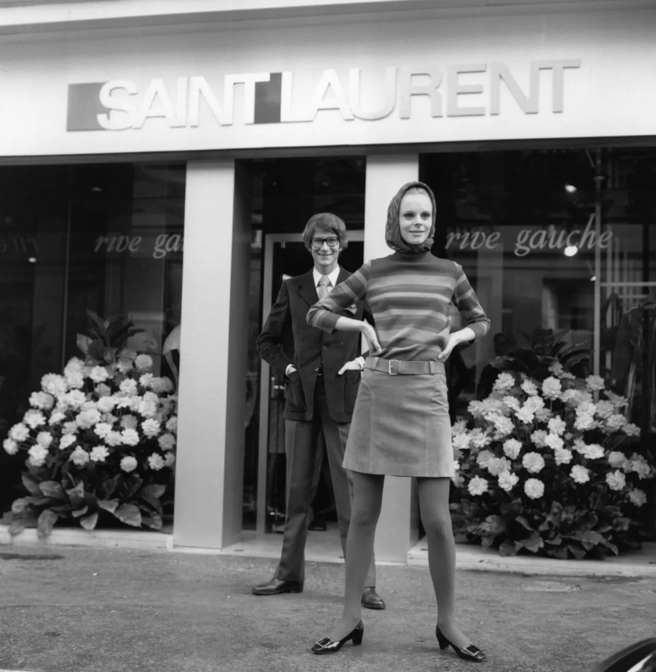 Yves St. Laurent standing outside his Rive Gauche boutique in France, with a model wearing an ensemble from his 1966 collection with “Belle Vivier” pumps.